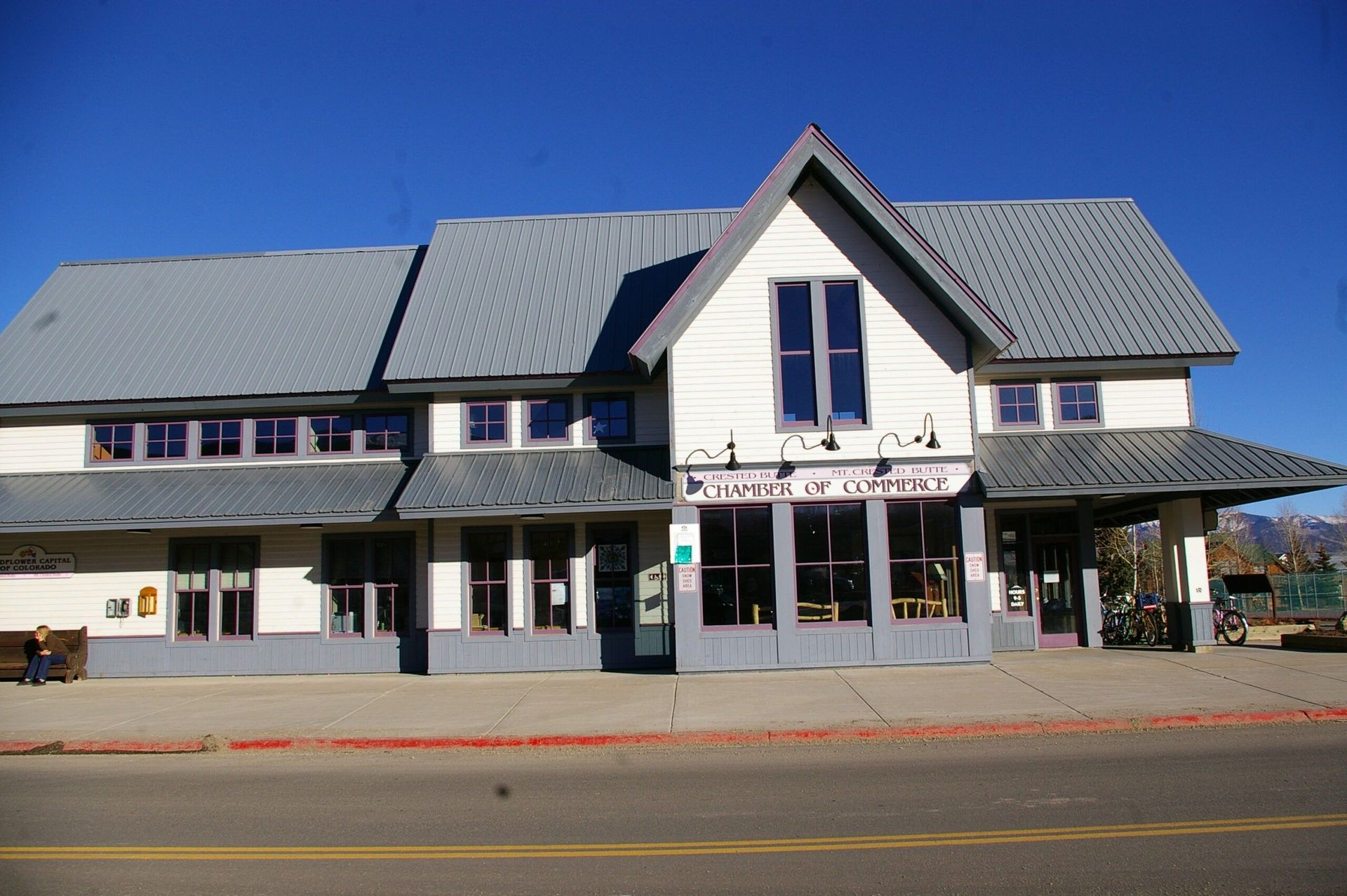 Two-story building with gray roof and white exterior, storefront with large windows, bright blue sky.