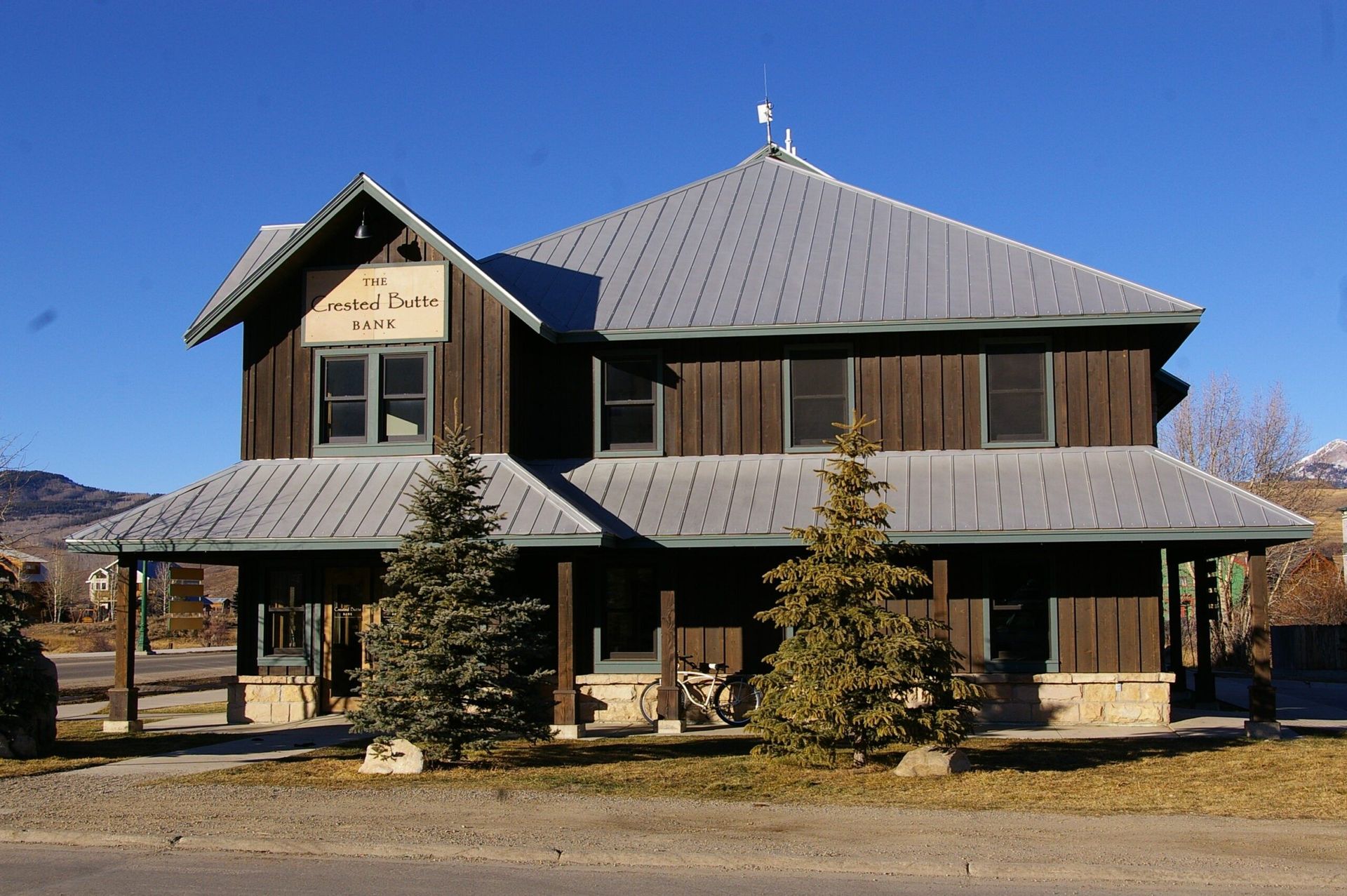Two-story brown building with gray roof and porch, evergreen trees in front, clear blue sky.