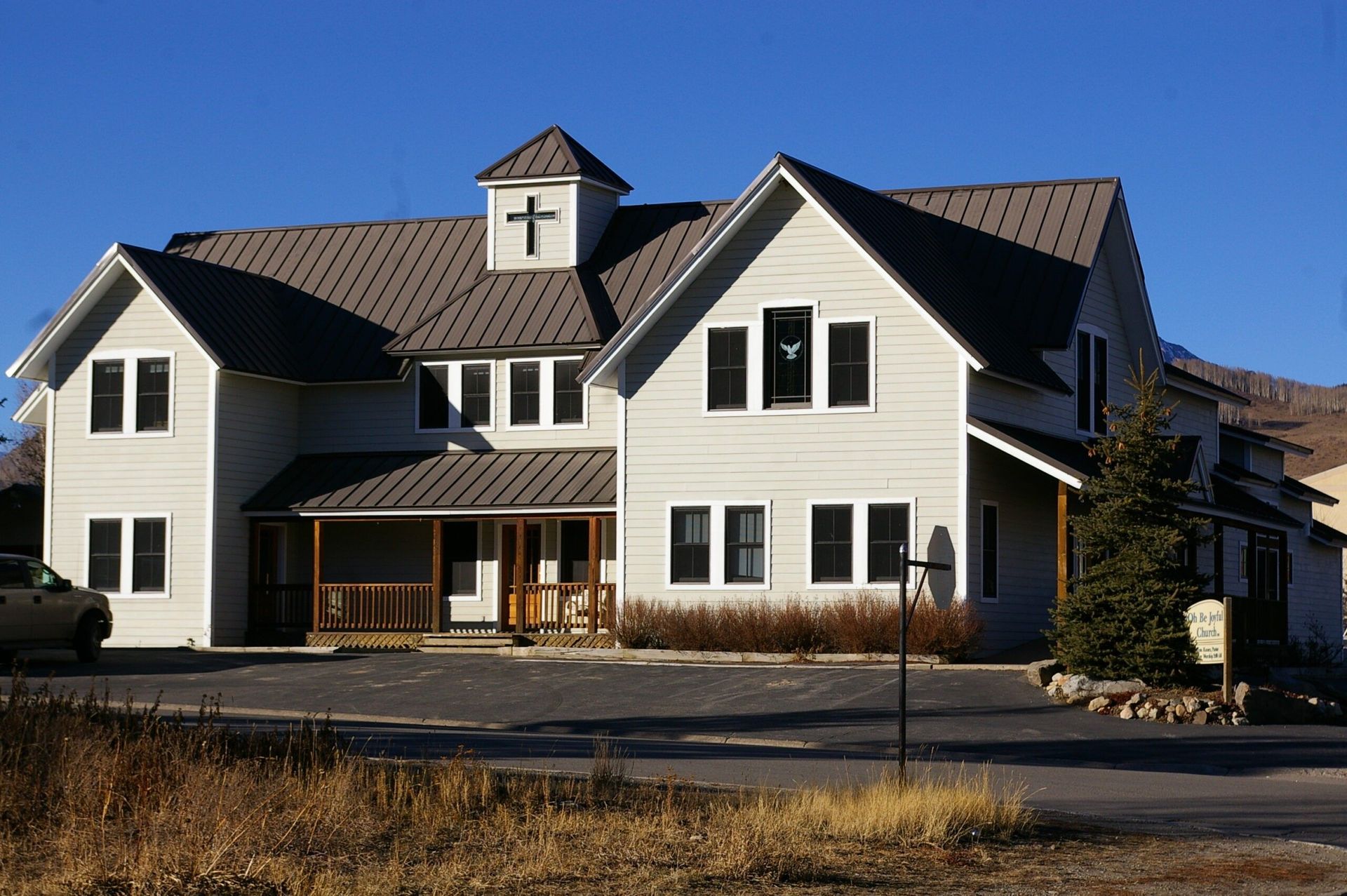 Two-story beige building with a metal roof, cross on top, in a sunny setting with a blue sky.