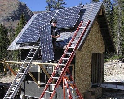 Man installing solar panels on a small cabin roof in a mountain setting.