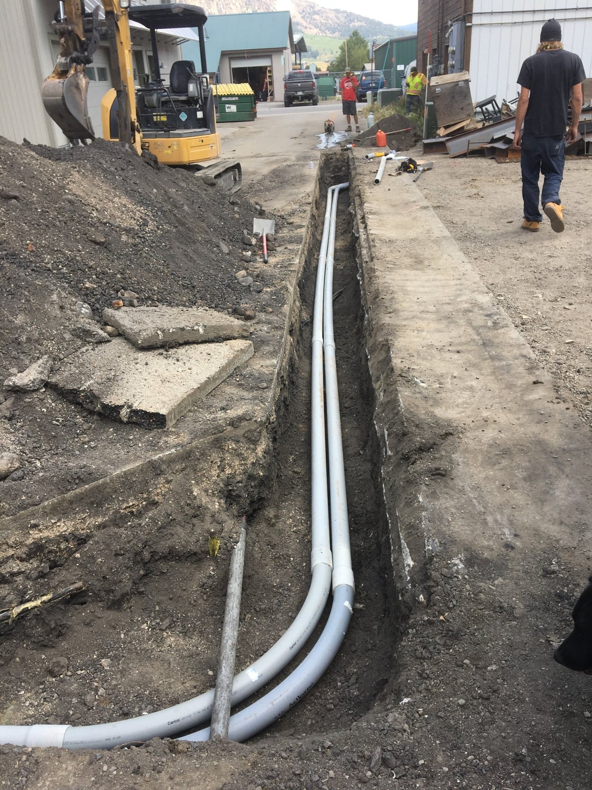 Construction site: grey electrical conduits in a trench, small excavator, workers, asphalt and dirt.