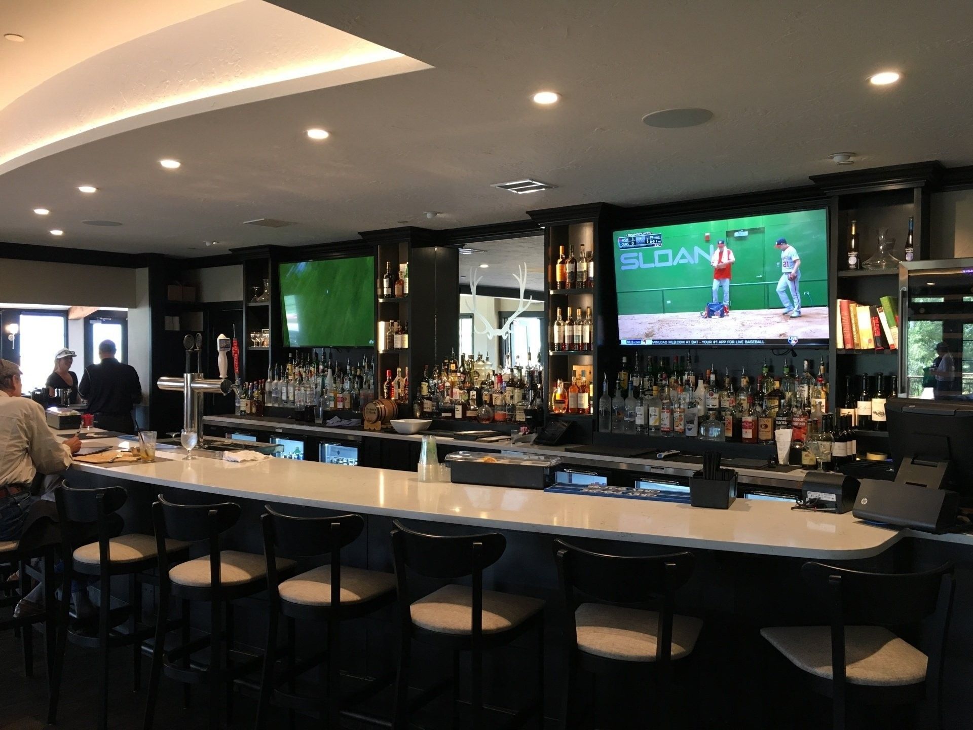 Bar with patrons seated, dark decor, large TV screens, and fully stocked shelves of alcohol.