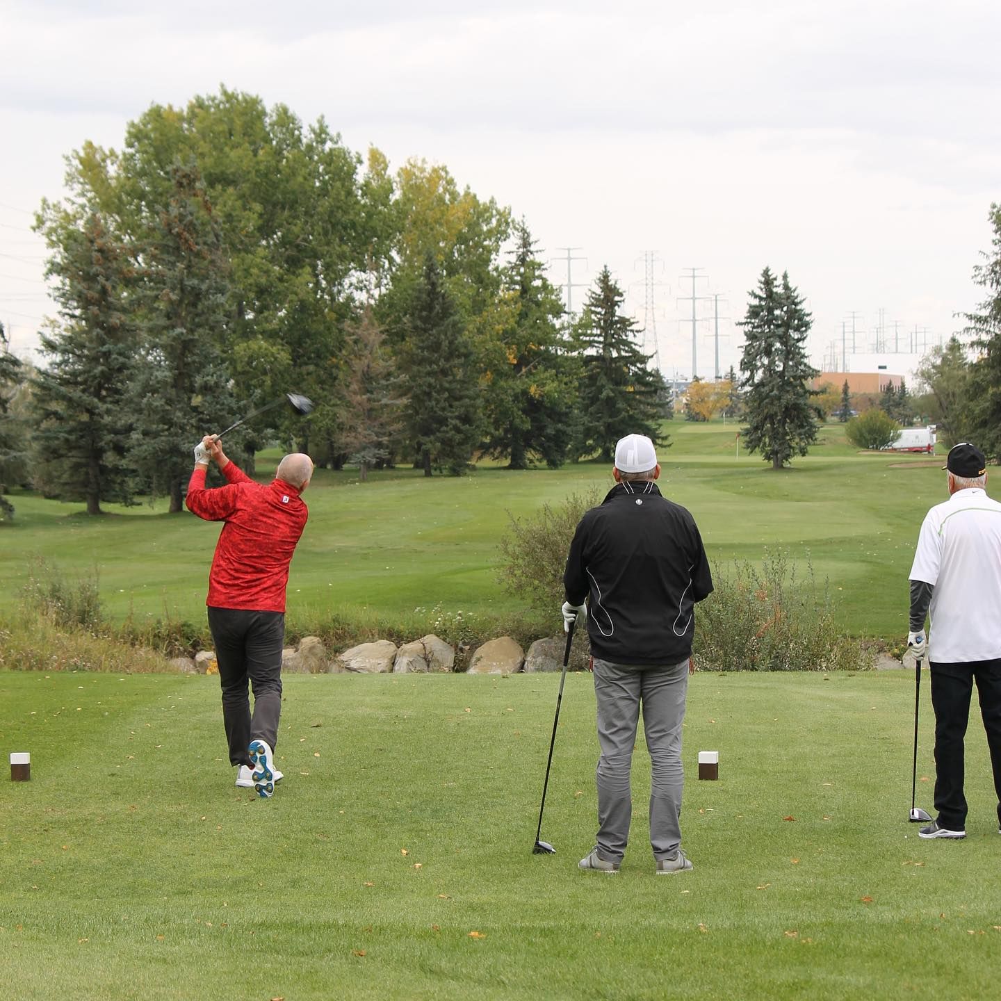 A golfer in red swings, watched by two men on a green golf course under an overcast sky.