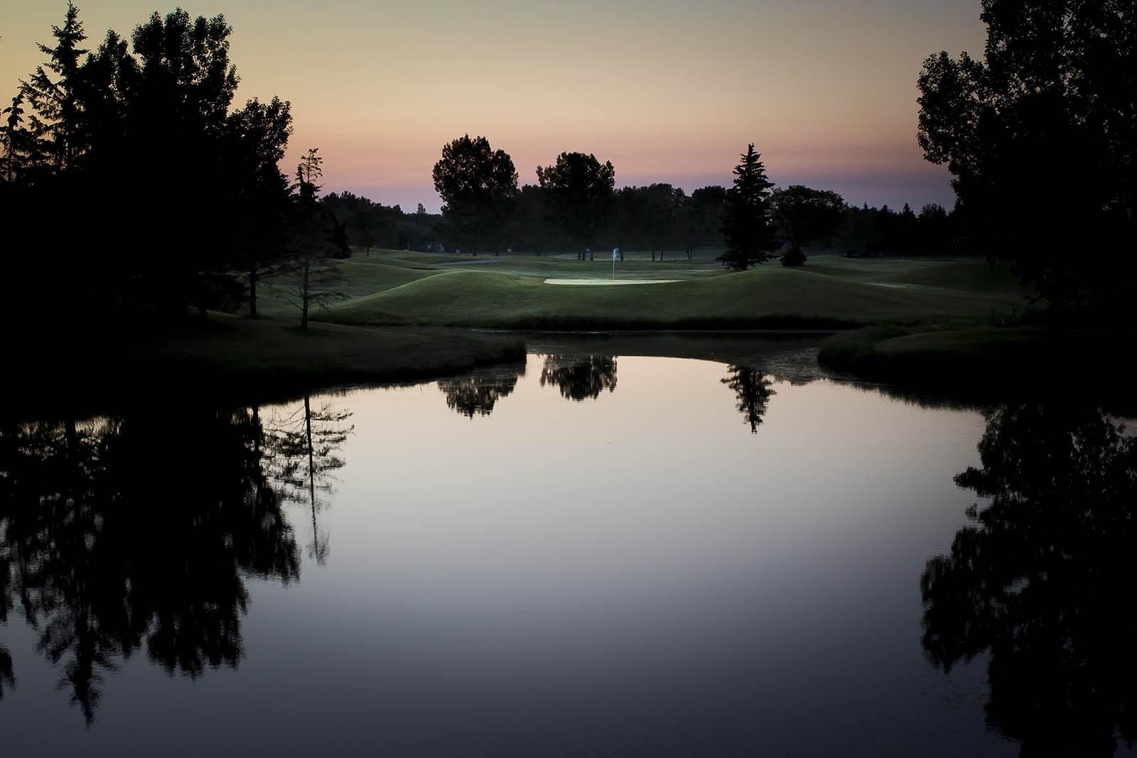 A serene golf course at dusk; trees reflected in calm water under a muted sky.