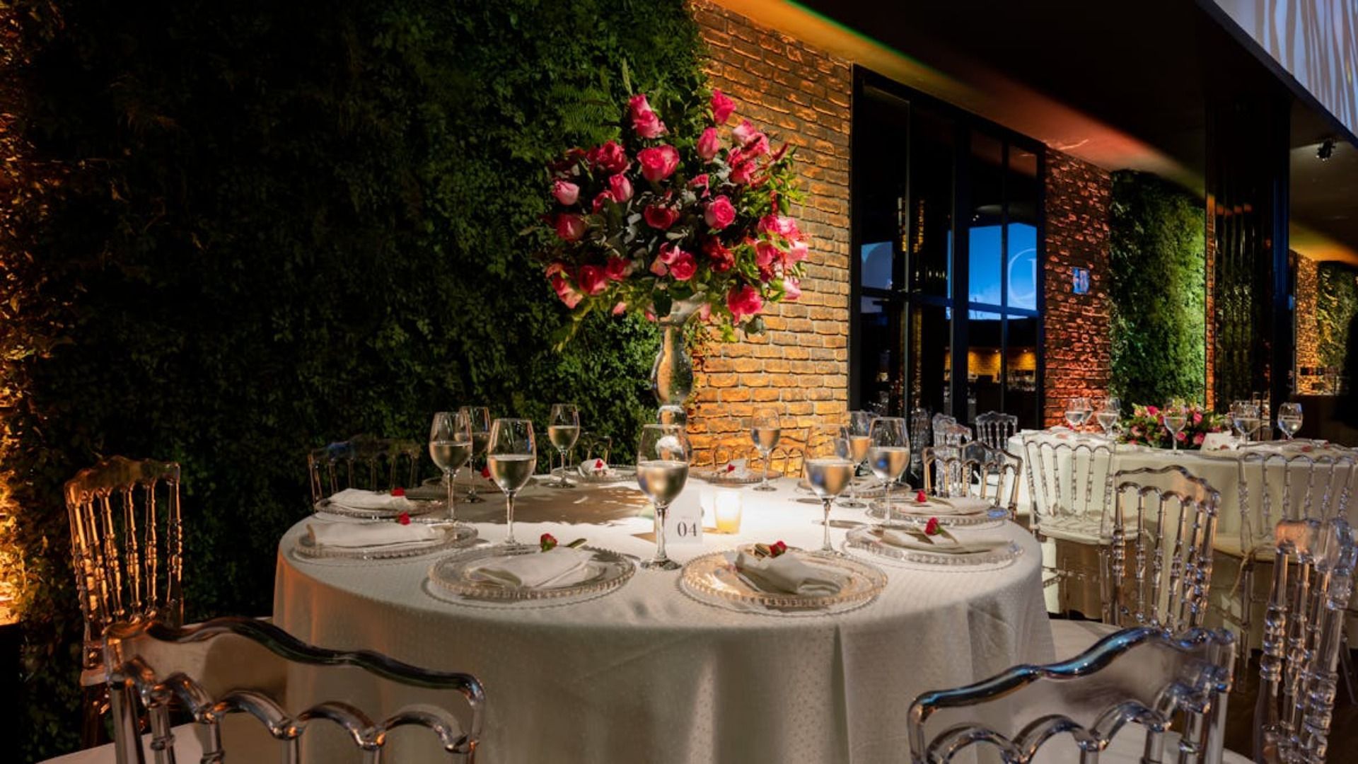 Una mesa redonda con manteles blancos y sillas transparentes en una habitación con luz tenue, elegantemente decorada