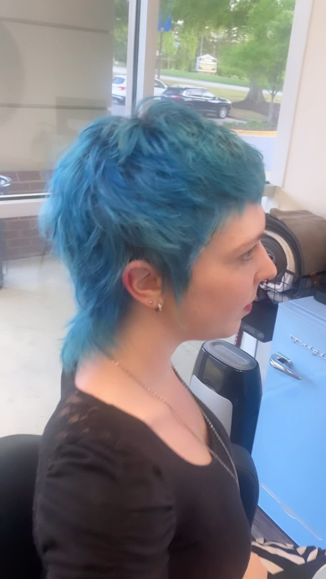 A woman with blue hair is sitting in a chair in front of a window.