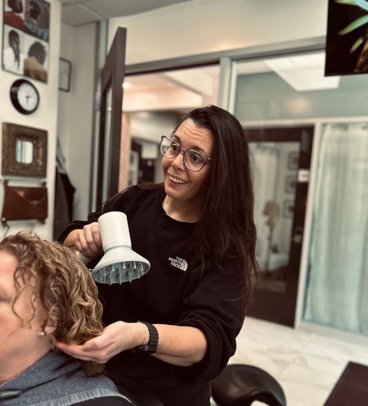 A woman with long blonde hair and a plaid shirt is standing in front of a wooden wall.