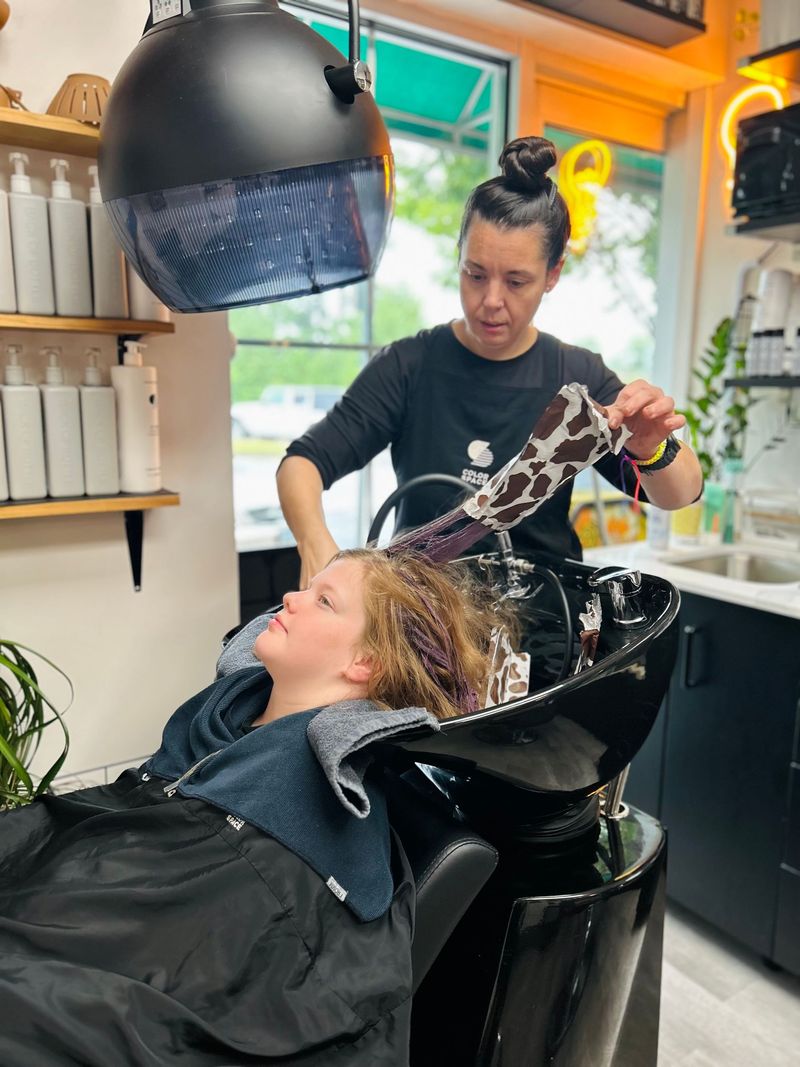 Woman washing hair of a child in a salon; black sink, woman in black shirt, overhead dryer, shelves with white bottles.