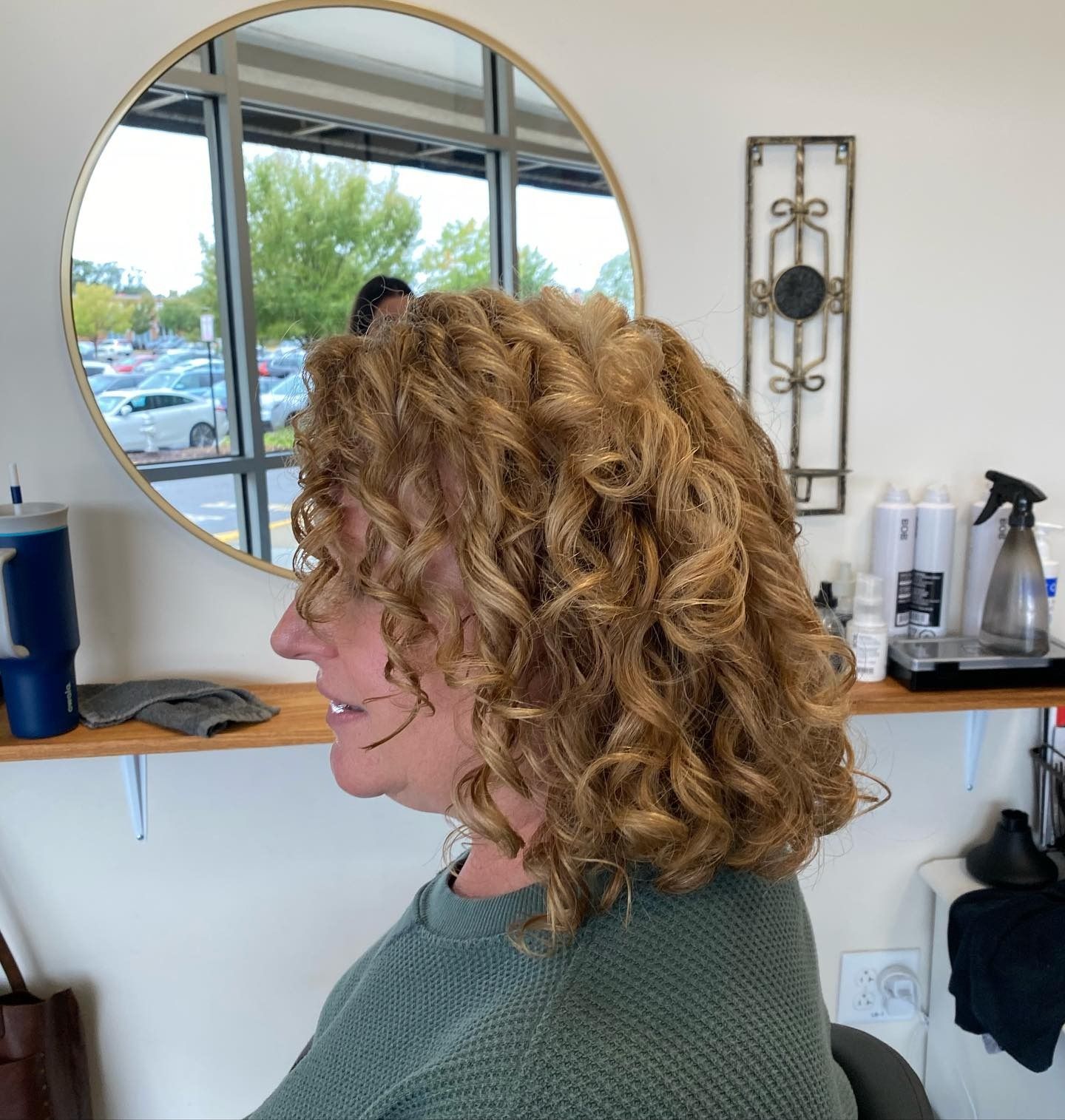 A woman with curly hair is sitting in front of a mirror in a salon.