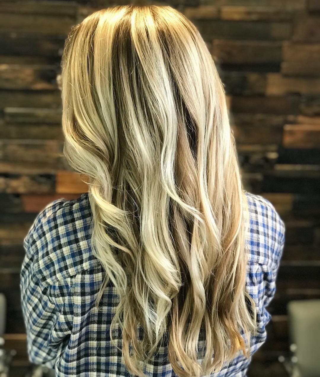 A woman with long blonde hair and a plaid shirt is standing in front of a wooden wall.