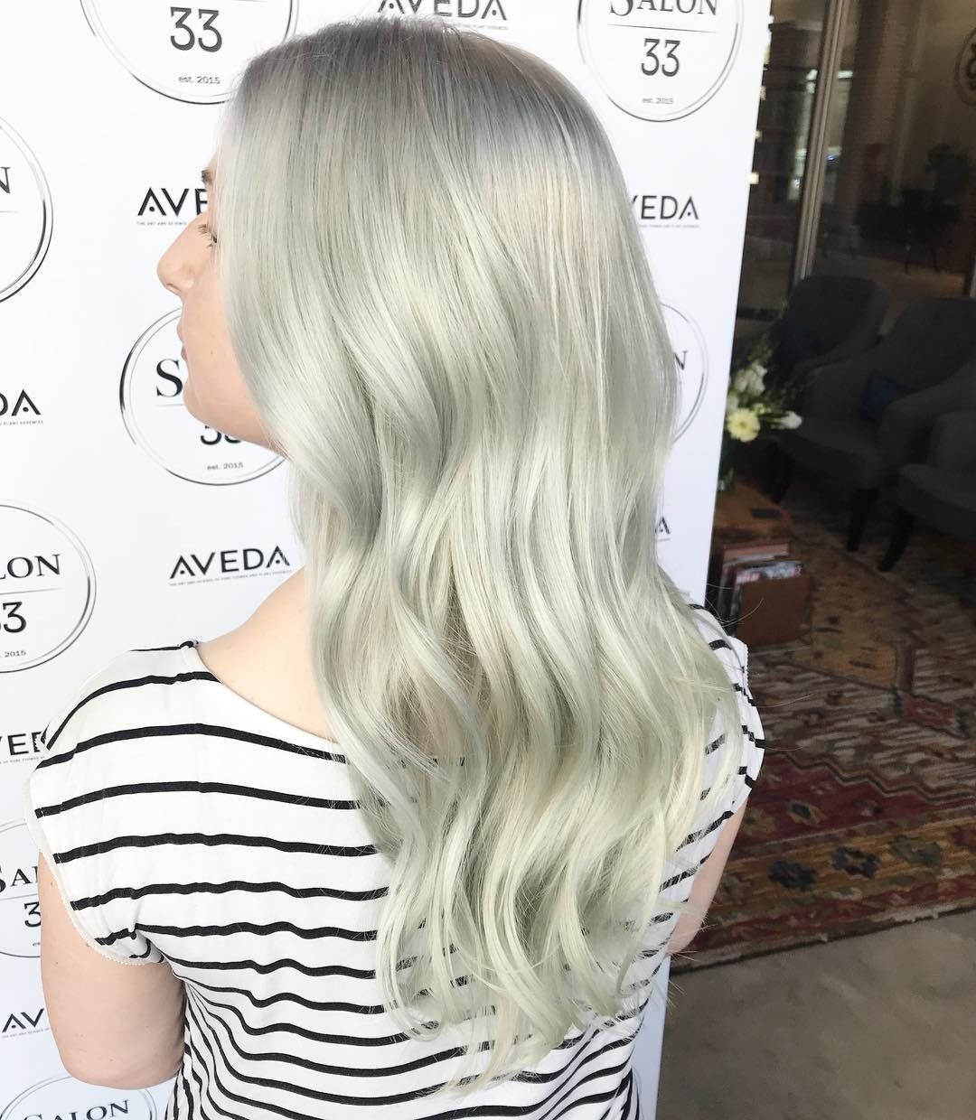 A woman with long white hair is standing in front of a sign that says aveda