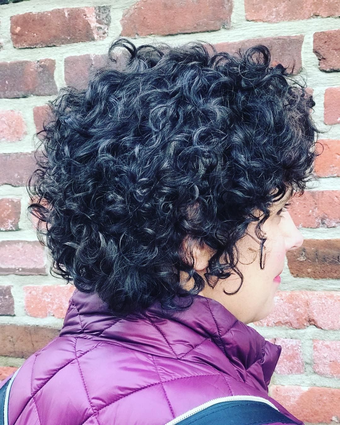 A woman with curly hair is wearing a purple jacket and standing in front of a brick wall.
