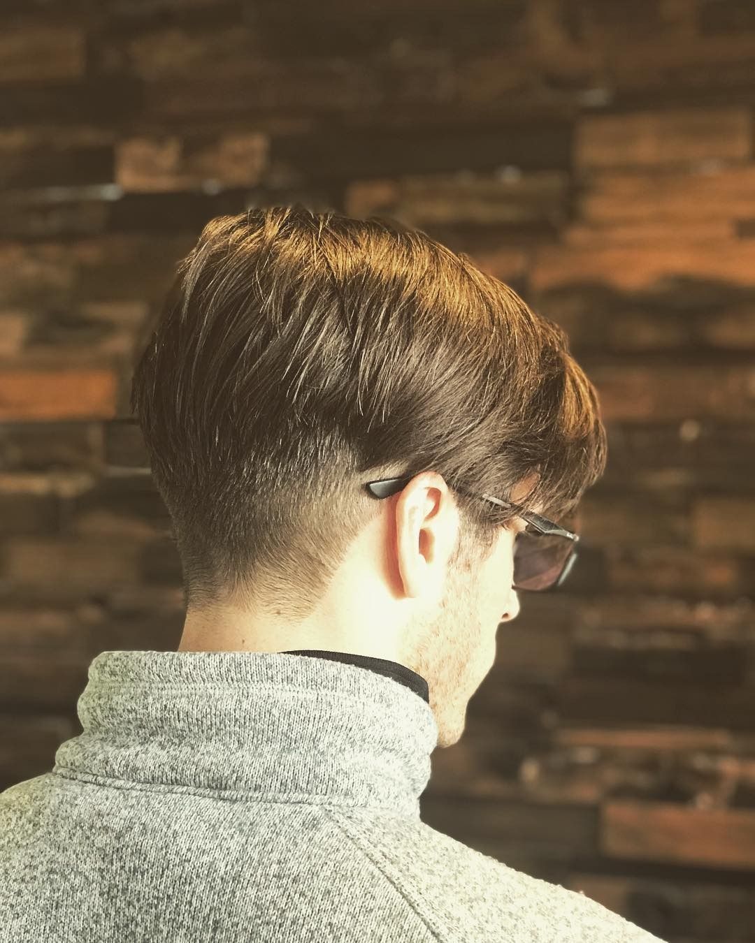 A man wearing sunglasses and a gray sweater is standing in front of a wooden wall.
