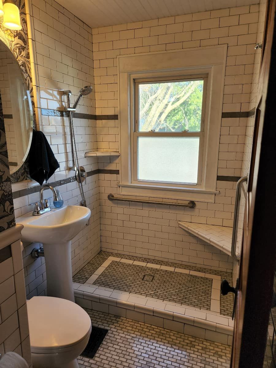 Small bathroom with white tiled walls, shower, pedestal sink, and window.