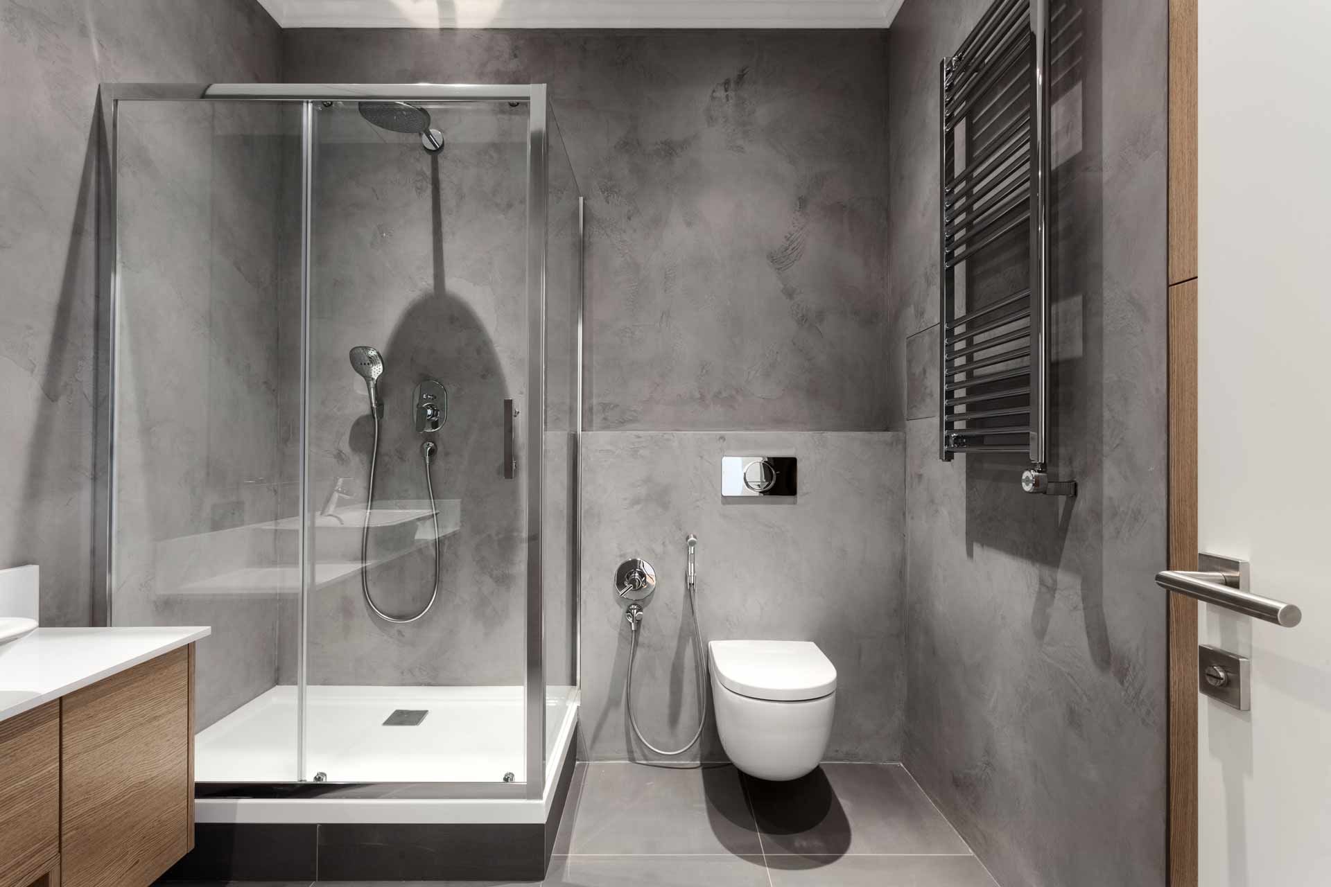 Modern bathroom with a glass shower, toilet, towel rack, and wooden cabinet. Grey walls and floor.