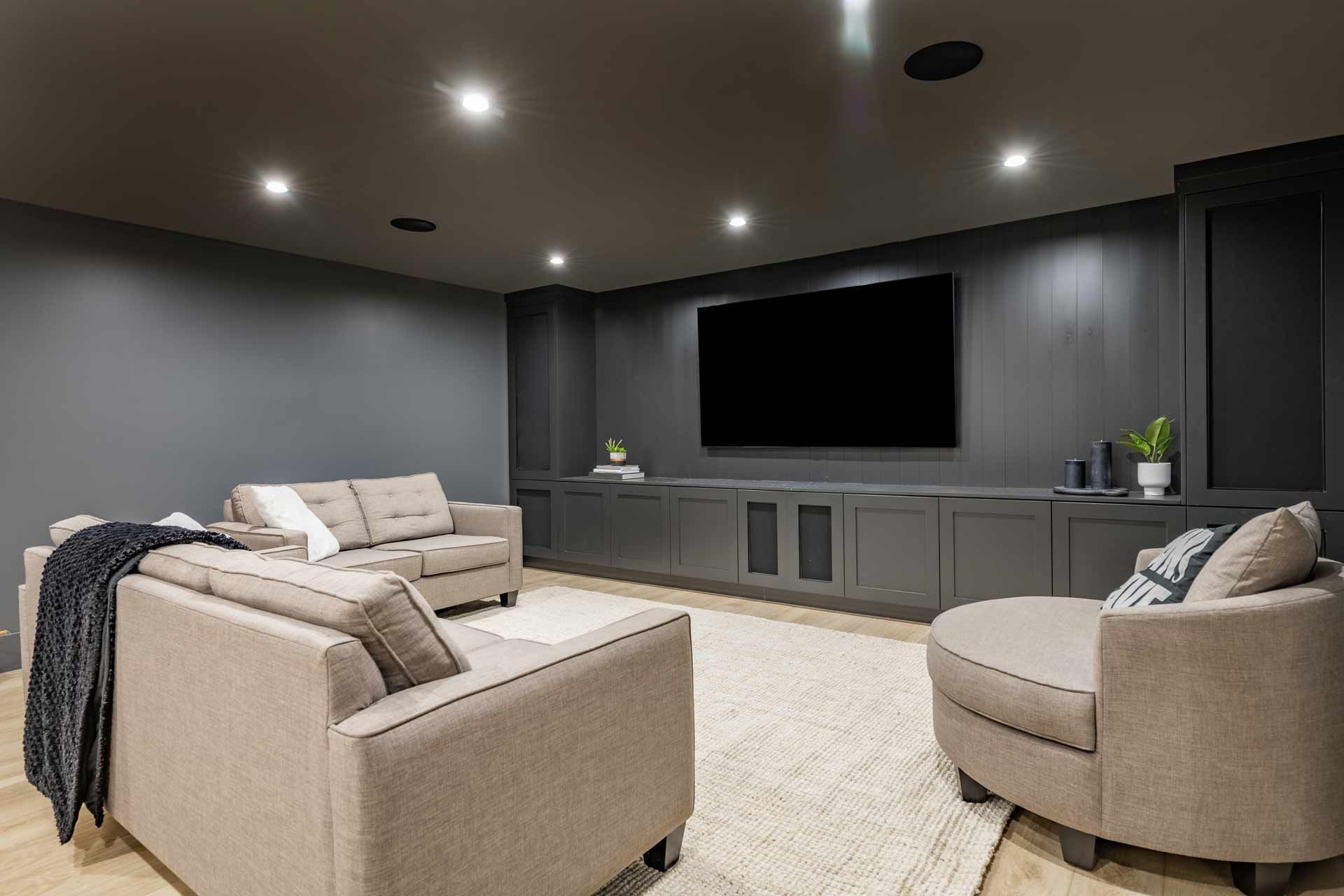 A dark gray home theater with neutral-toned furniture, large TV, and recessed lighting.