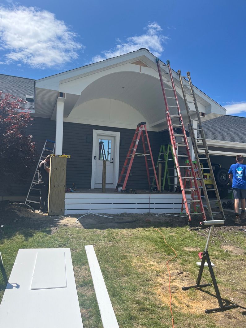 House porch under construction with ladders, workers, and materials on a sunny day.