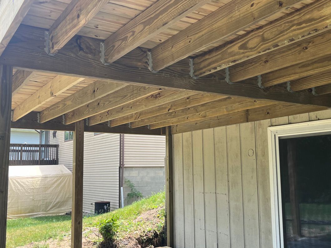 Underside of a wooden deck, featuring support beams and planks, overlooking a grassy area and a house.