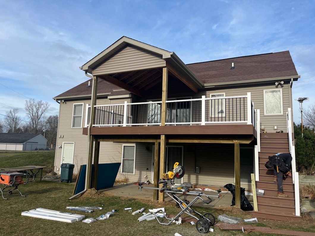 House with wooden deck under construction, person working on stairs, tools and materials scattered on the grass.