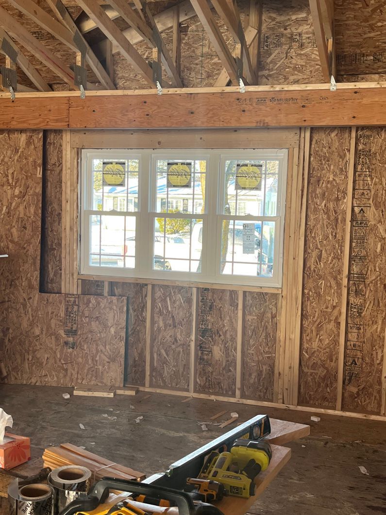 Interior framing of a building with a triple-pane window. OSB walls, wood beams, and construction tools are visible.