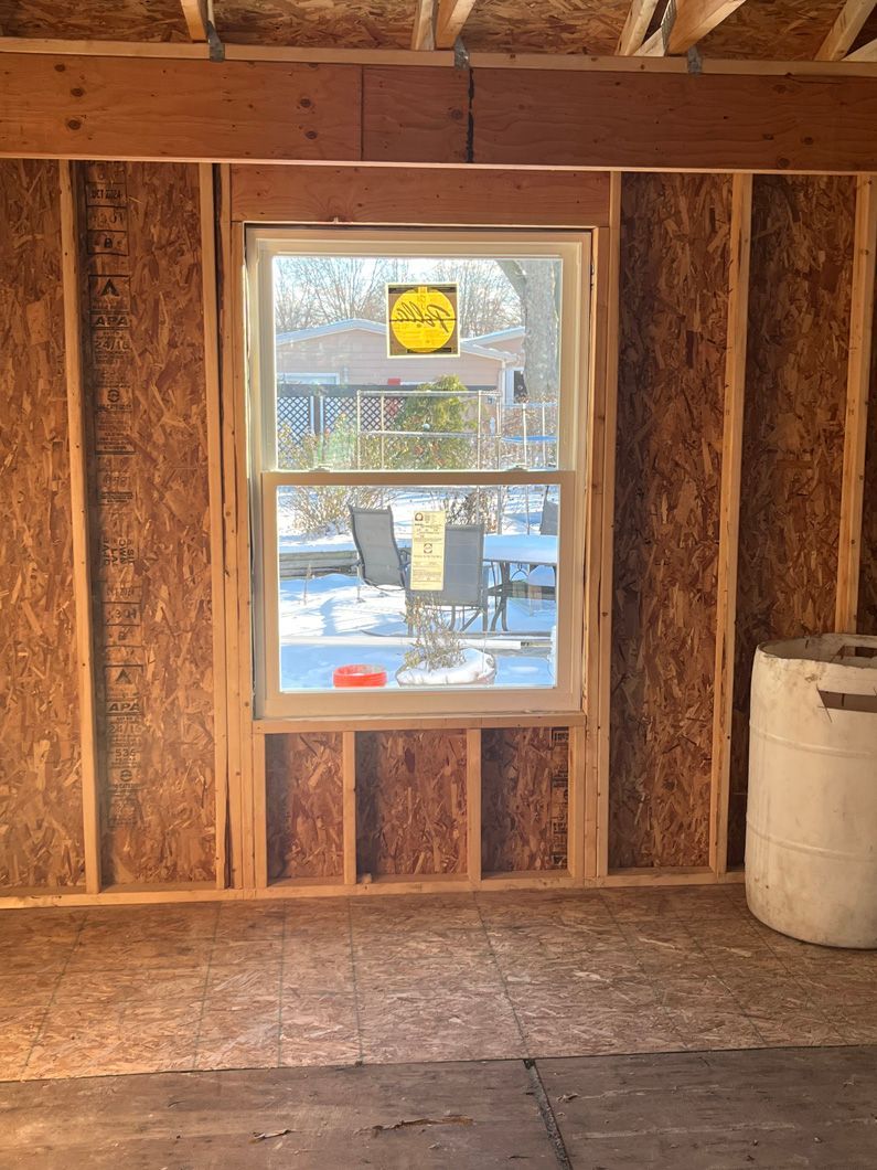 Interior of a room under construction, with a window, wood frame, and OSB walls, looking out to a yard.