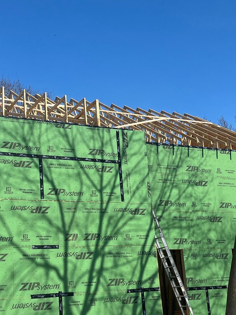 Construction site with green wall wrap, wood rafters against a blue sky.