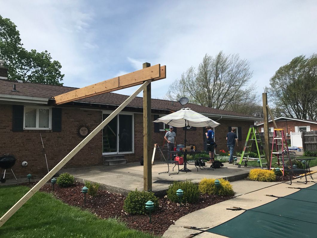 Construction of a patio structure. Workers assemble beams and support posts next to a pool, yard, and house.