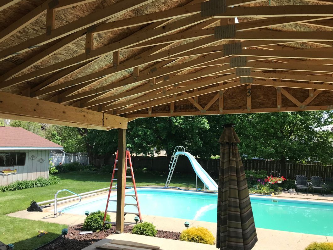 Wooden patio cover construction over a backyard pool. A-frame rafters, ladder, and umbrella visible.