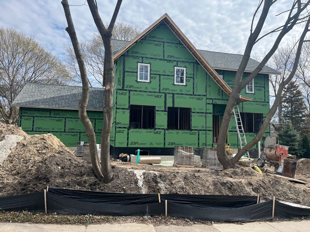 A two-story house under construction, covered in green wrap. Dirt and trees in the foreground.