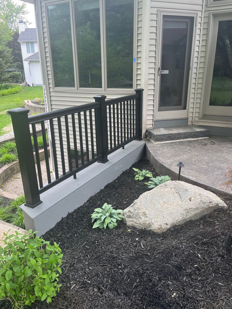Black railing on gray wall near a stone, with plants and windows in the background.