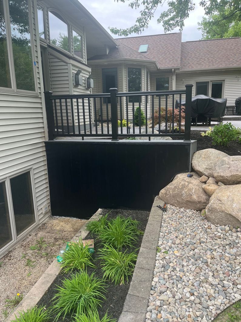 Black deck with black railing against a beige house; garden with rocks and greenery in foreground.