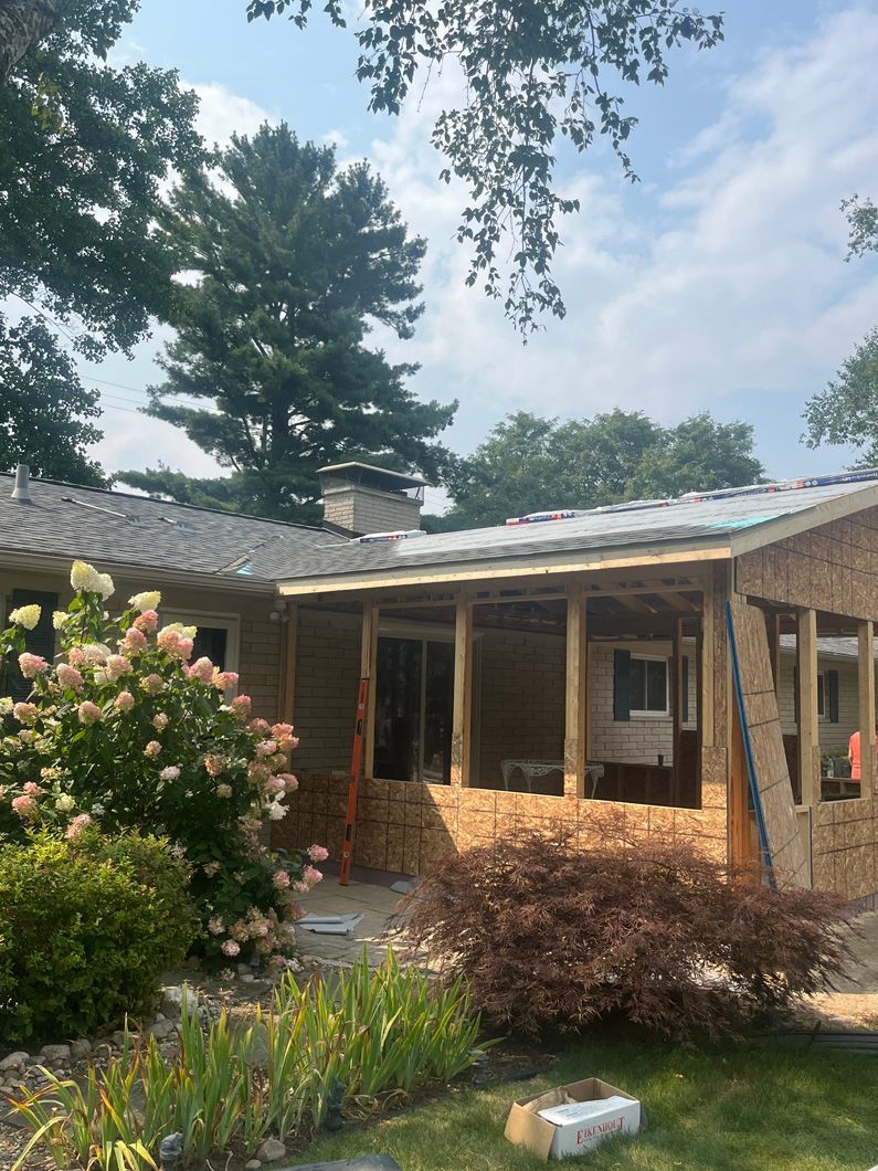 House exterior under construction with exposed framing and brickwork, trees and foliage in the foreground.