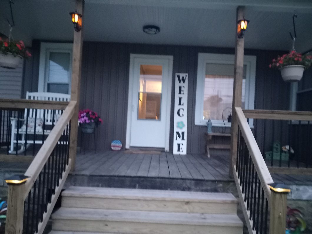 Front porch with wooden steps and railing. A welcome sign stands by the white door.