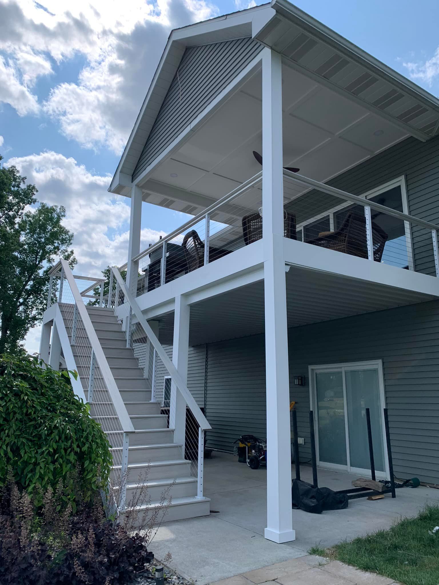Two-story deck with stairs, white railings, and a screened roof, attached to a gray house on a sunny day.