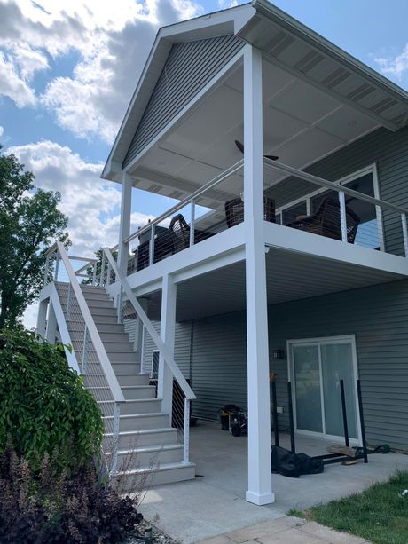 Two-story deck with stairs, white railings, and a screened roof, attached to a gray house on a sunny day.