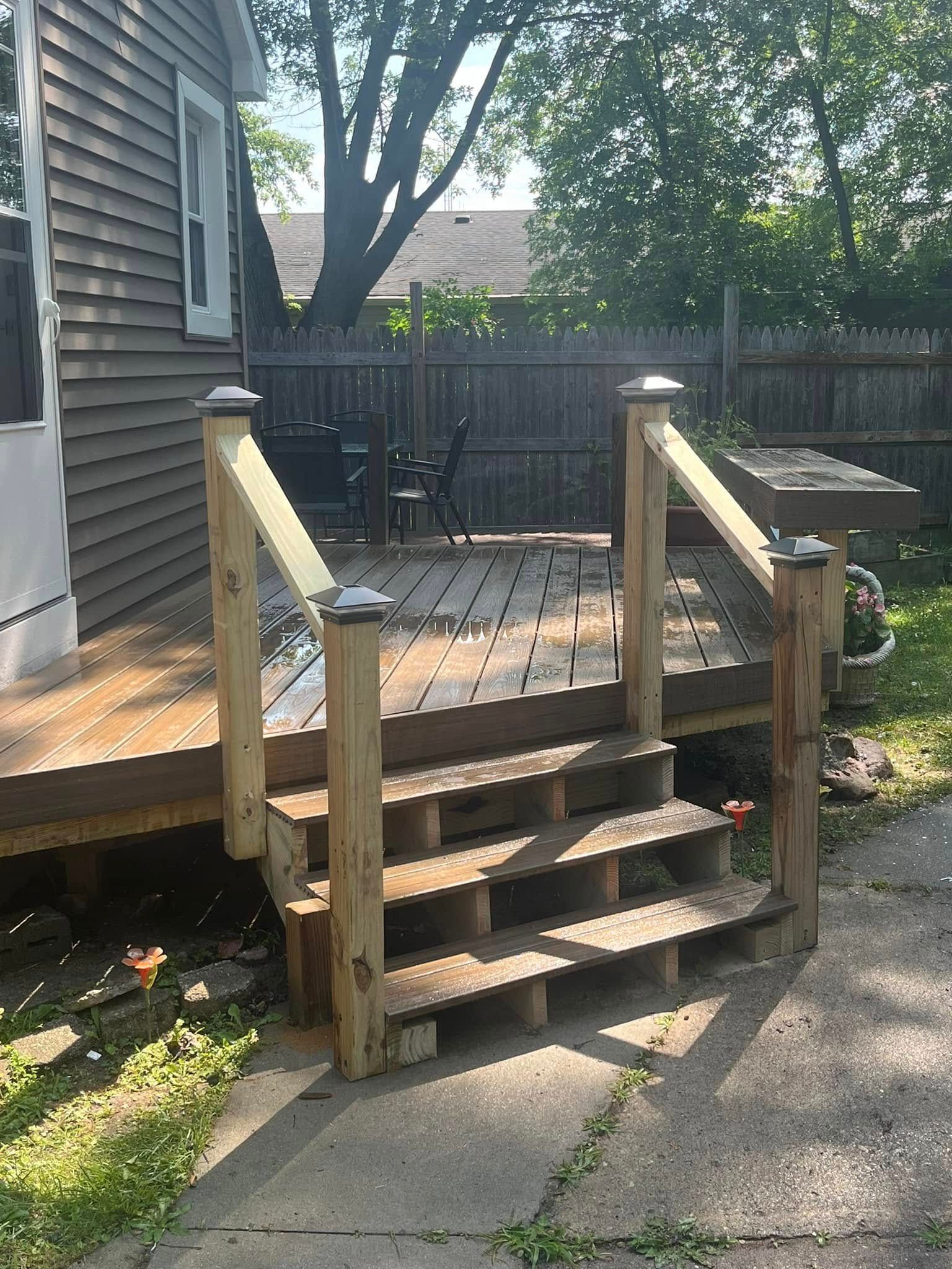 Wooden deck with steps, railing, and post caps. House exterior in background.