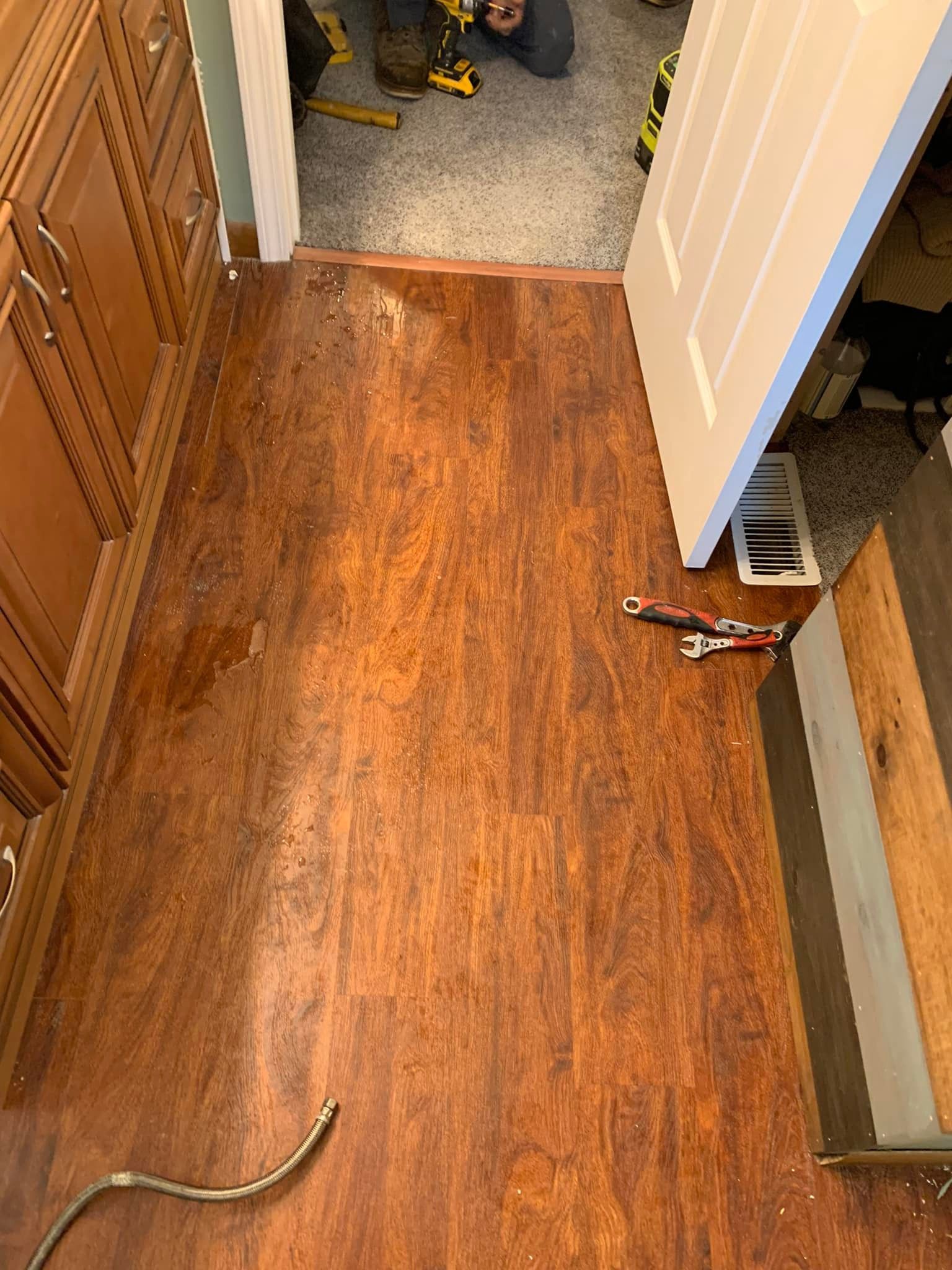 Laminate flooring being installed in a room, tools scattered nearby. Cabinet and open doorway visible.