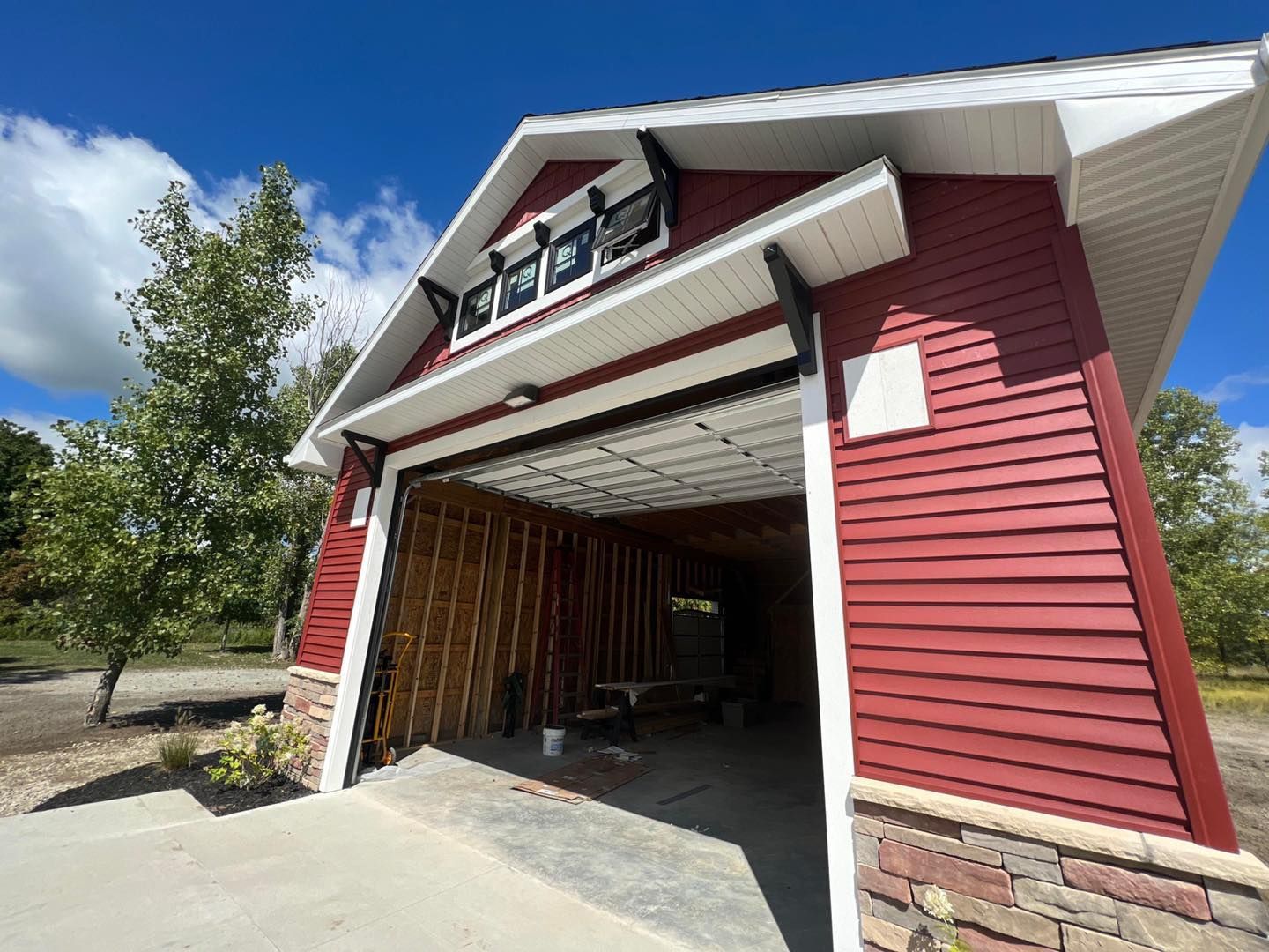 Red barn-style garage with open door; brown accents, white trim, and stone base. Bright blue sky.