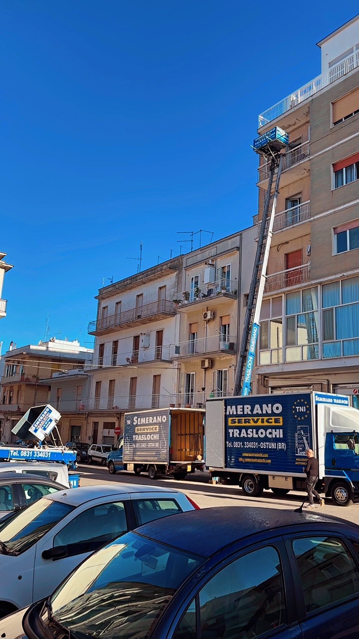Camion per traslochi che scarica mobili in un edificio, con un elevatore esteso; cielo azzurro.