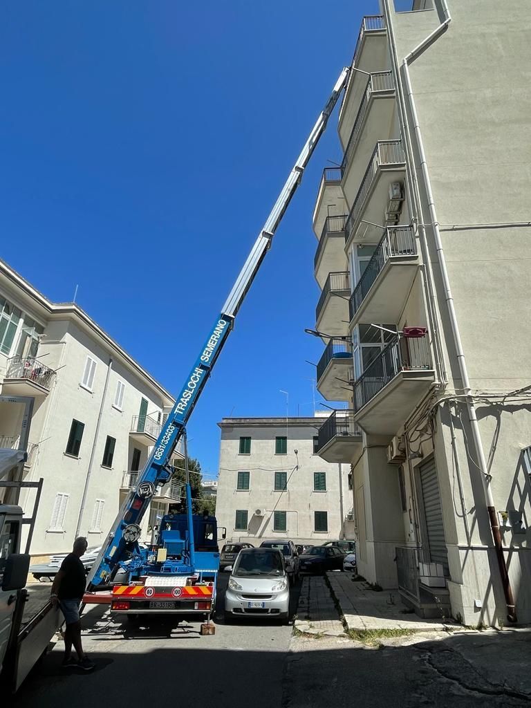 Gru blu che si estende verso un edificio a più piani per lo spostamento. Uomo in piedi vicino a un camion. Giornata di sole splendente.
