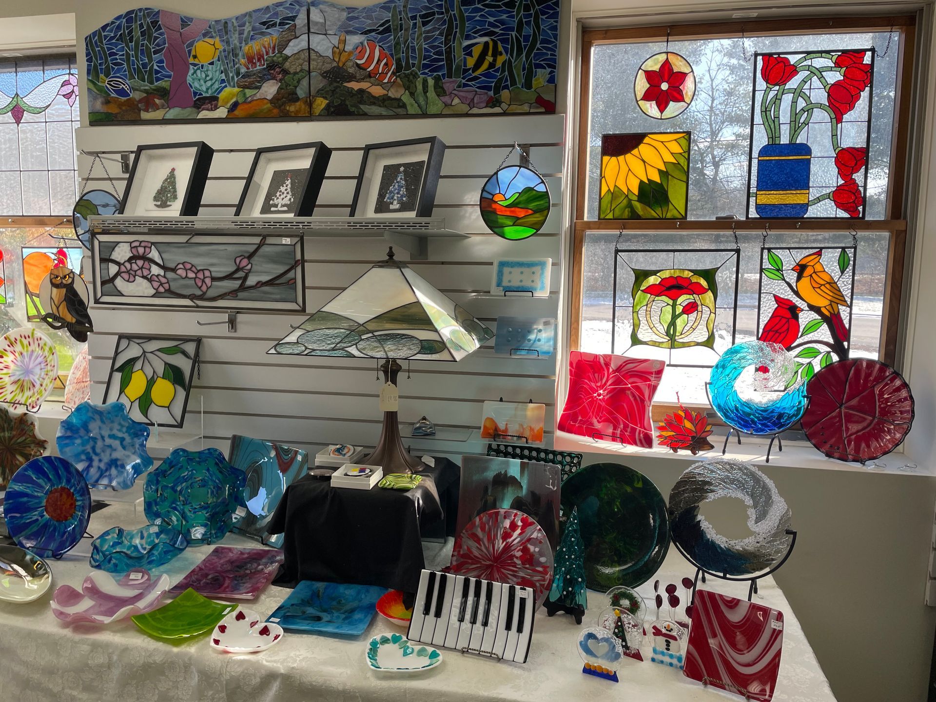 Glass art display: stained glass panels, plates, bowls, and other items on a white table and shelves.