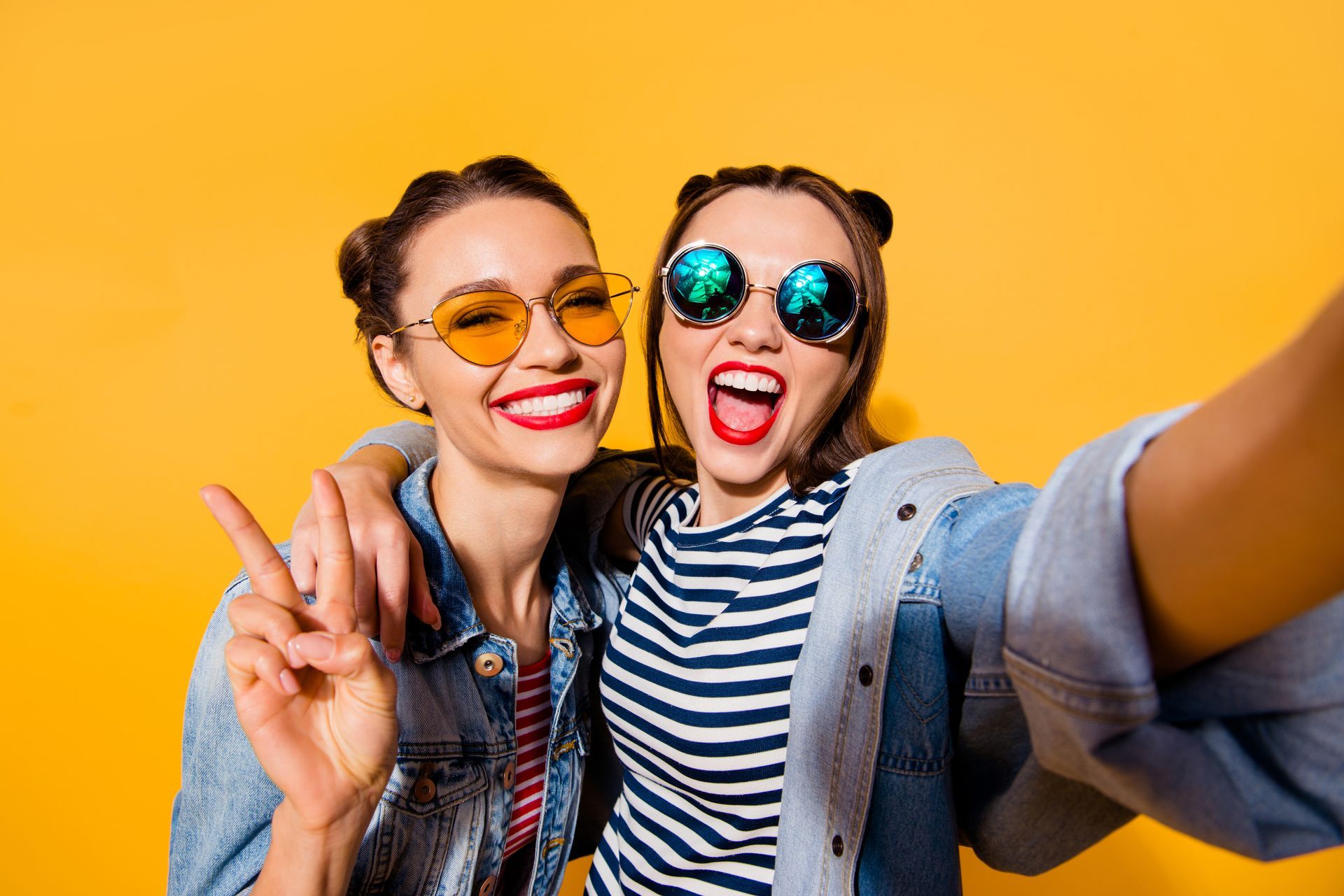 Two women in sunglasses smiling, taking a selfie against a yellow background. One gives a peace sign.