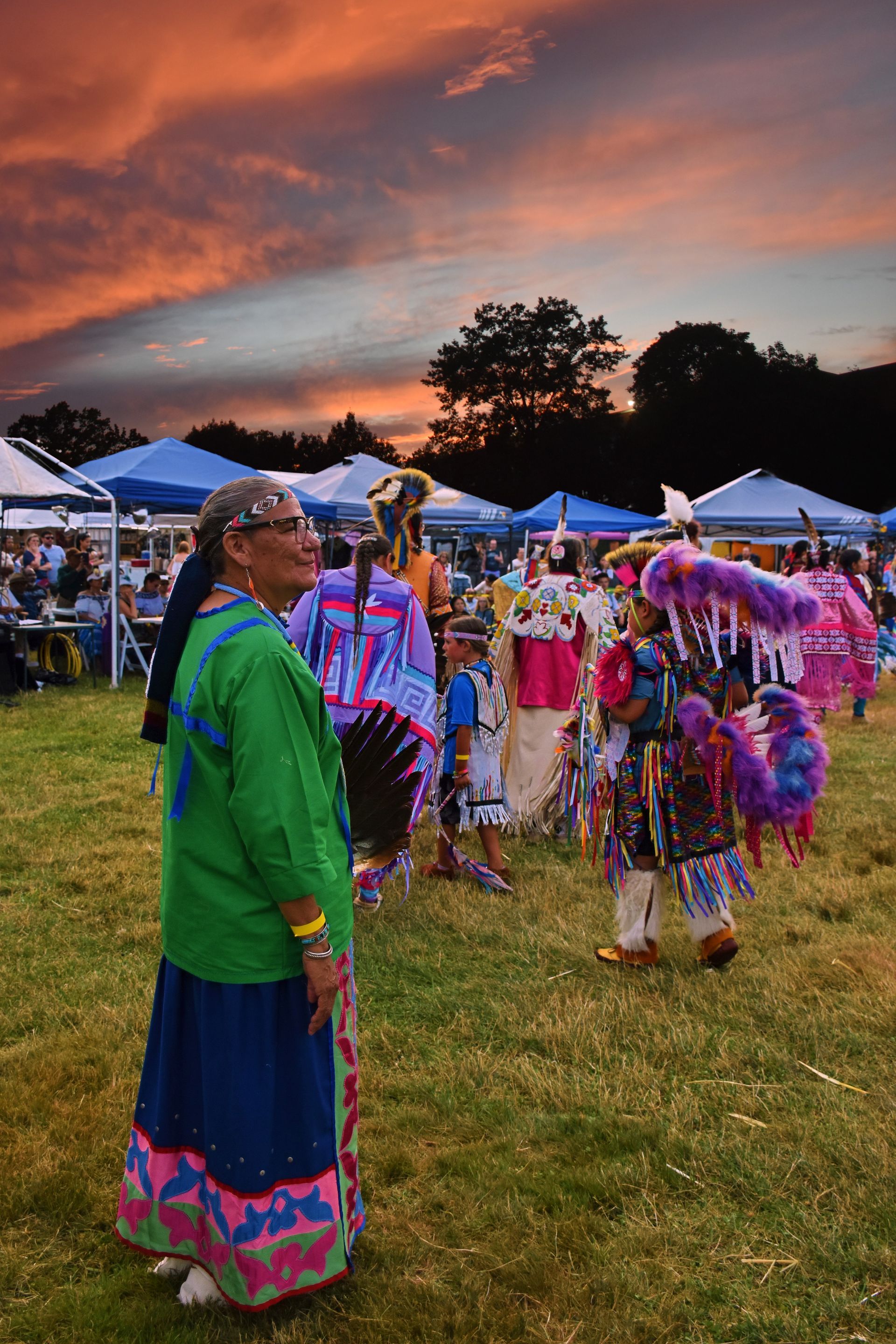 Woman in green, watching dancers in Native American regalia at outdoor event; sunset background.