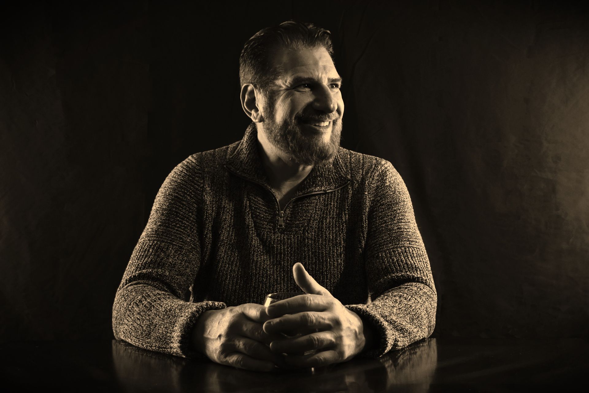 Man with a beard in a sweater smiles and clasps hands on a reflective surface against a dark background.