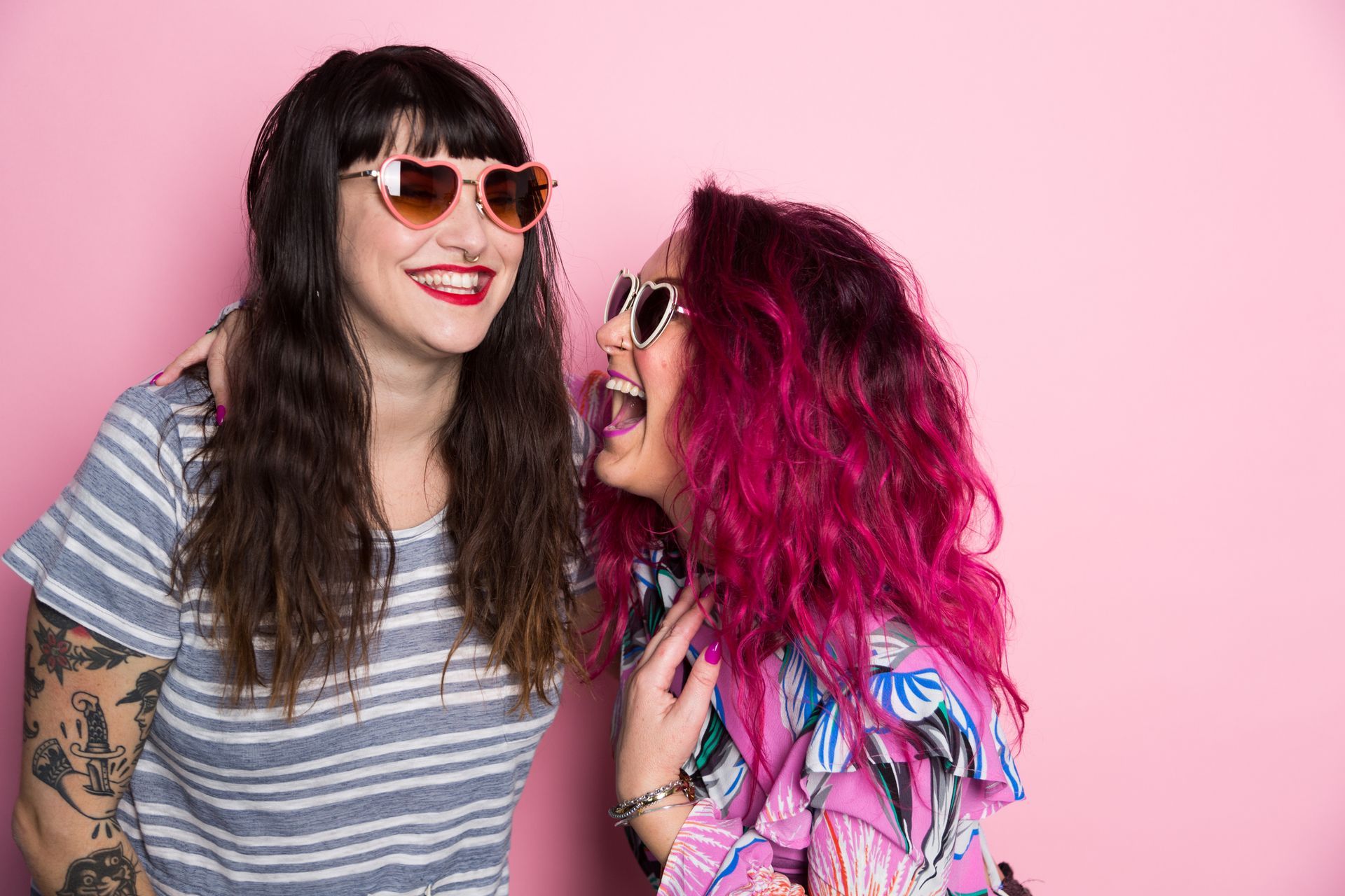 Two women wearing sunglasses laugh against a pink backdrop. One has dark hair, the other has pink hair.