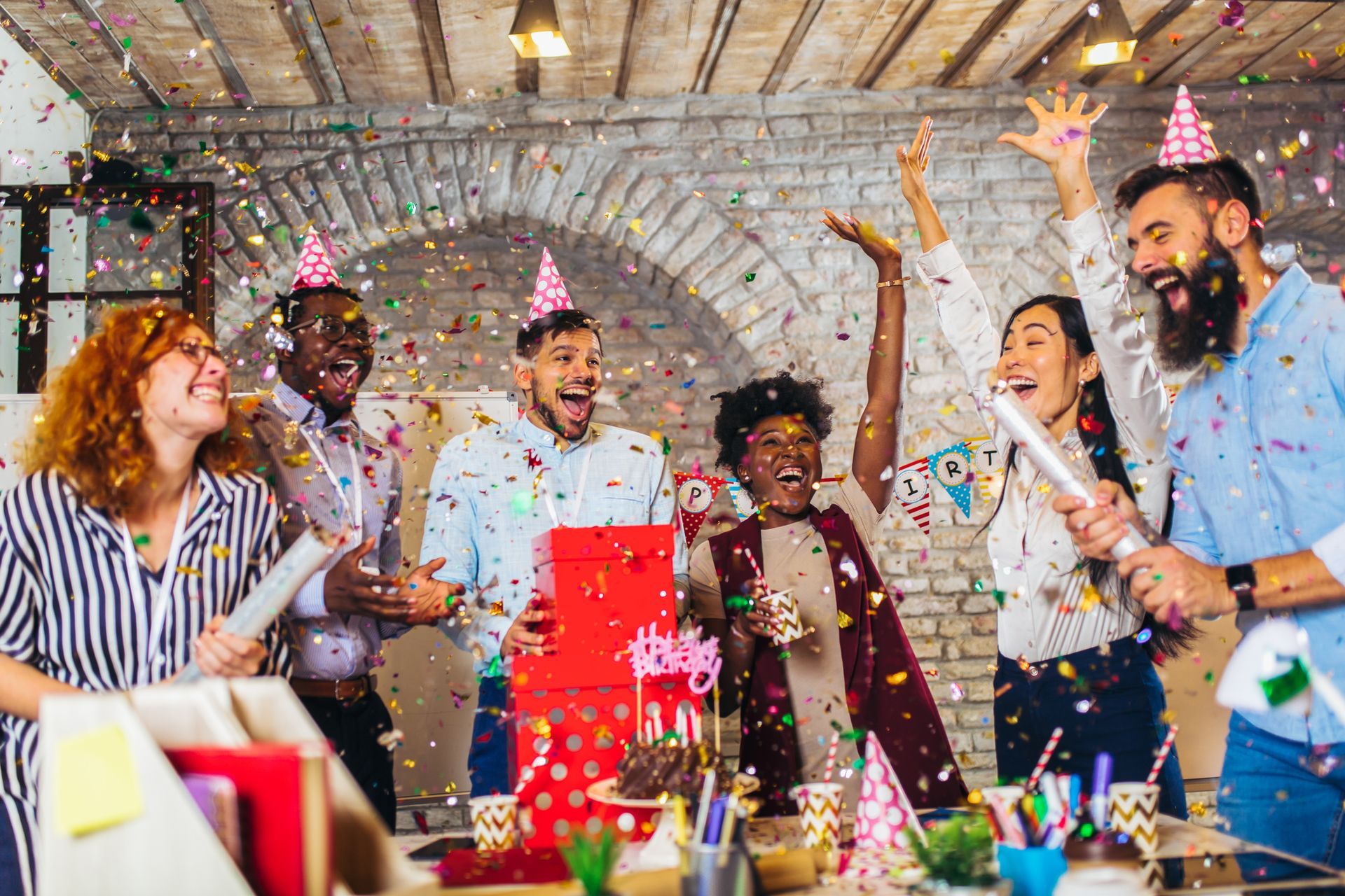 People in party hats celebrating with confetti and gifts in an office setting.