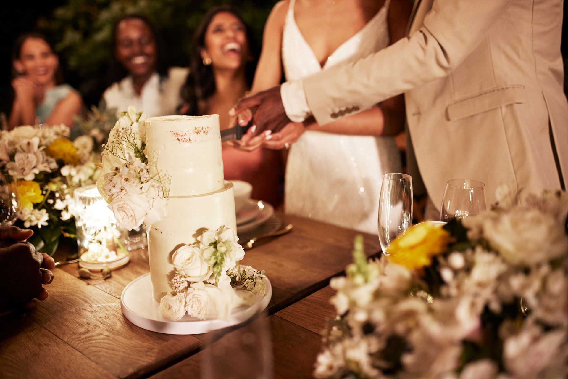 Wedding cake cutting; couple, friends, table set with flowers, laughing.