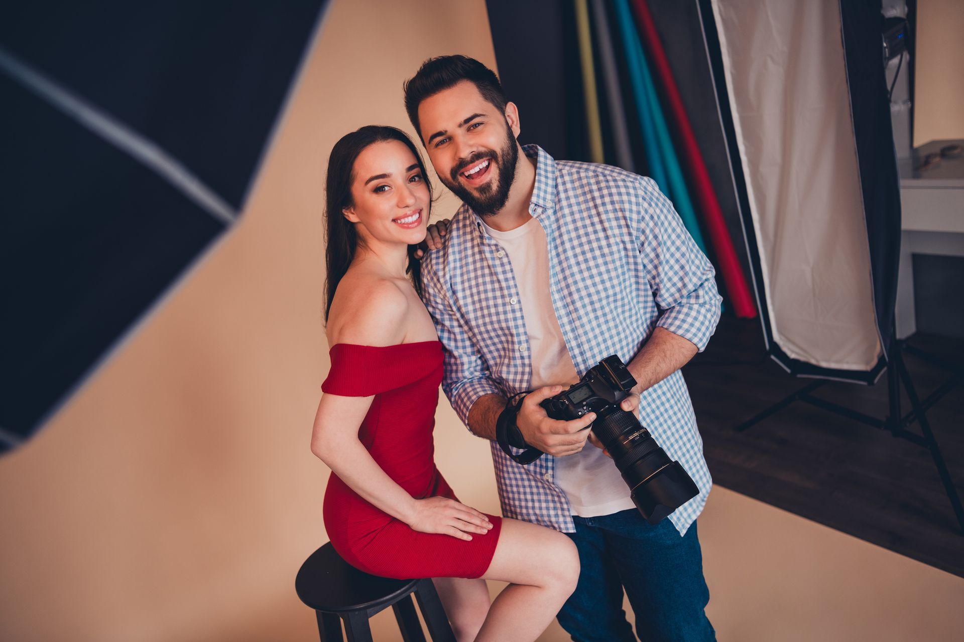 Woman in red dress sits with man holding a camera; both smile at the camera in a studio setting.