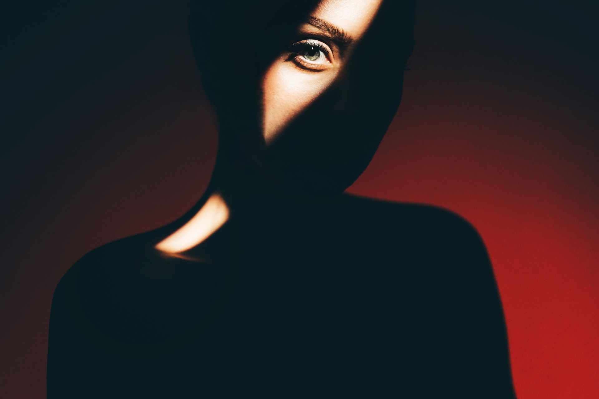 Person's face in shadow, one eye illuminated by light. Red background.
