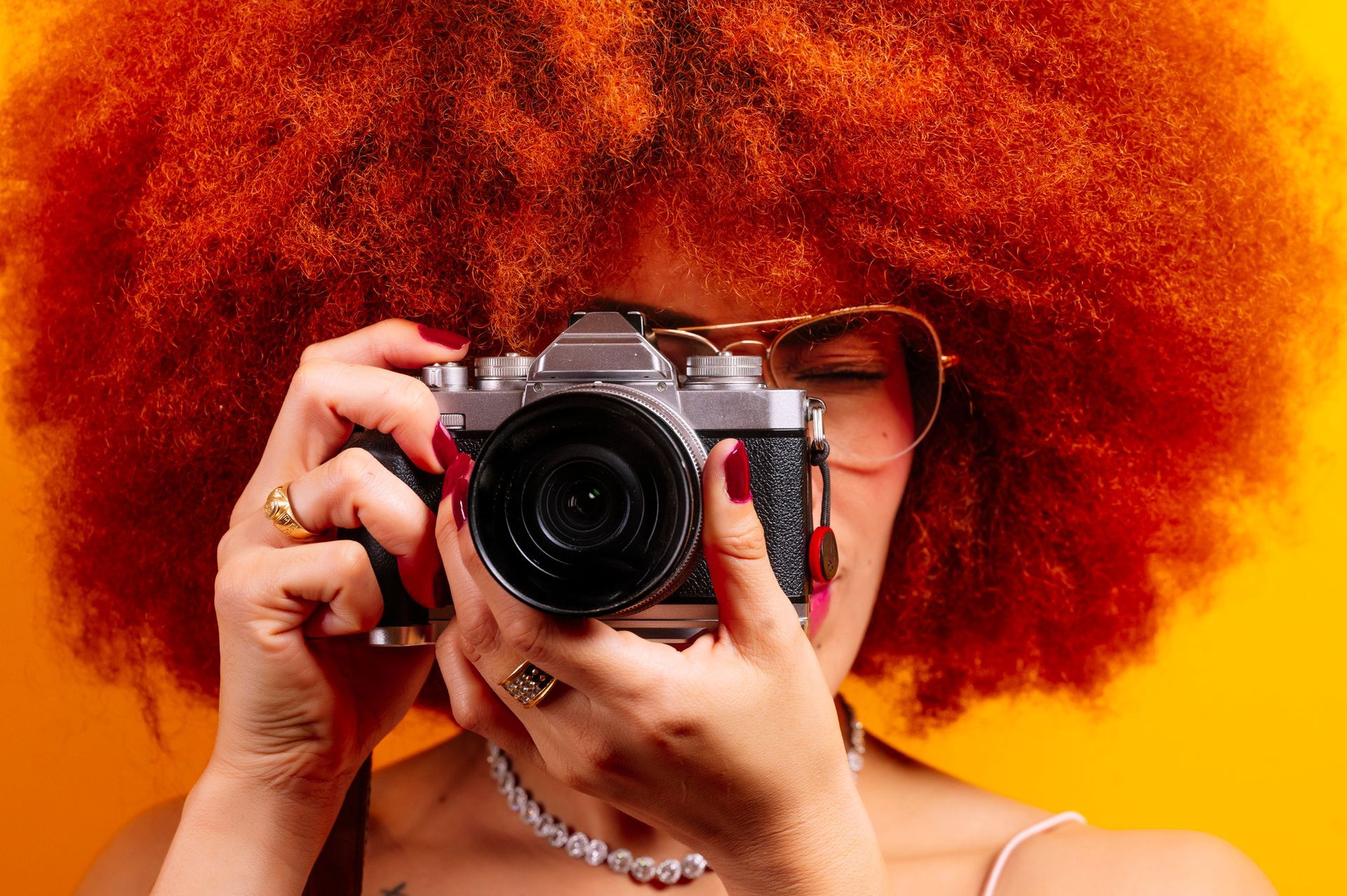 Woman with bright orange afro holding a vintage camera, wearing sunglasses and pearl necklace, against a yellow backdrop.